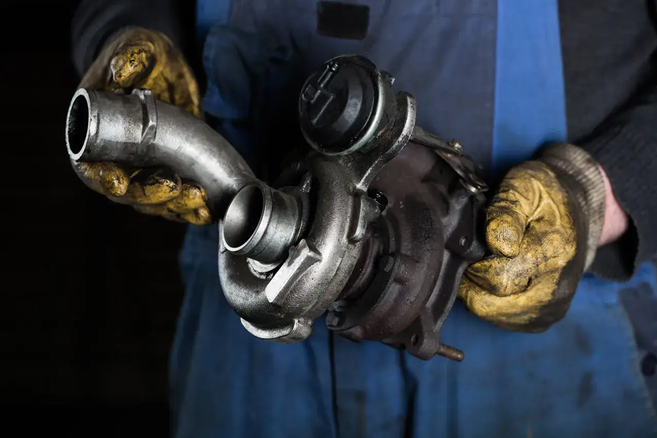 Mechanik w żółtych rękawicach trzyma turbosprężarkę. Objawy przeładowania turbiny mogą być różne, warto sprawdzić jej stan.