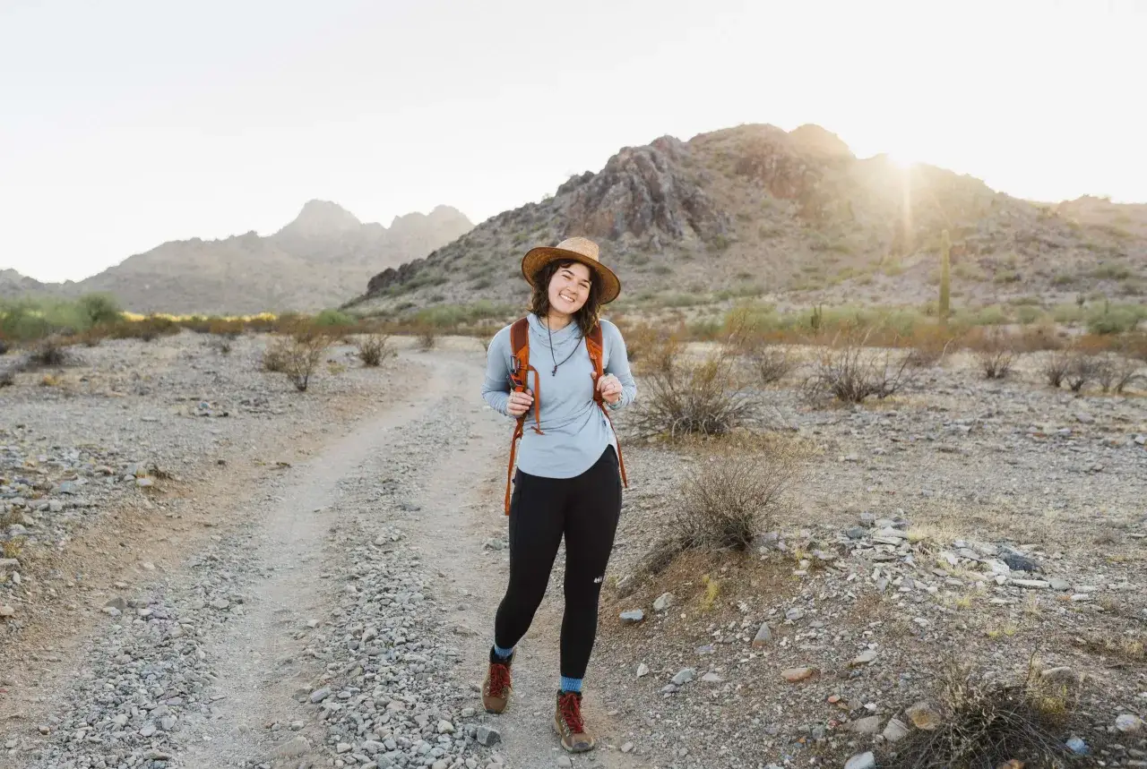 Donna sorridente con zaino e cappello di paglia su un sentiero desertico, un esempio di come vestirsi in montagna con abbigliamento comodo e funzionale.