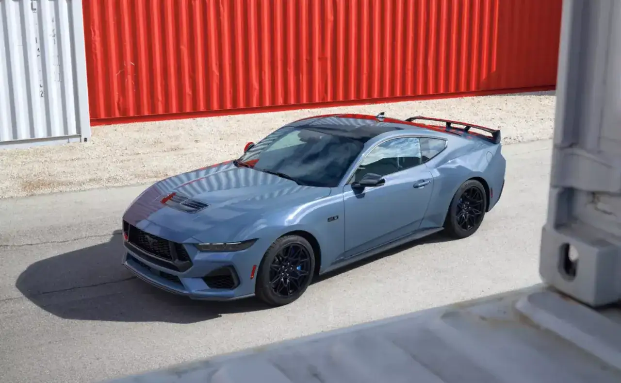 Niebieski Mustang 2026 z czarnym dachem i spoilerem, zaparkowany przy czerwonych kontenerach.