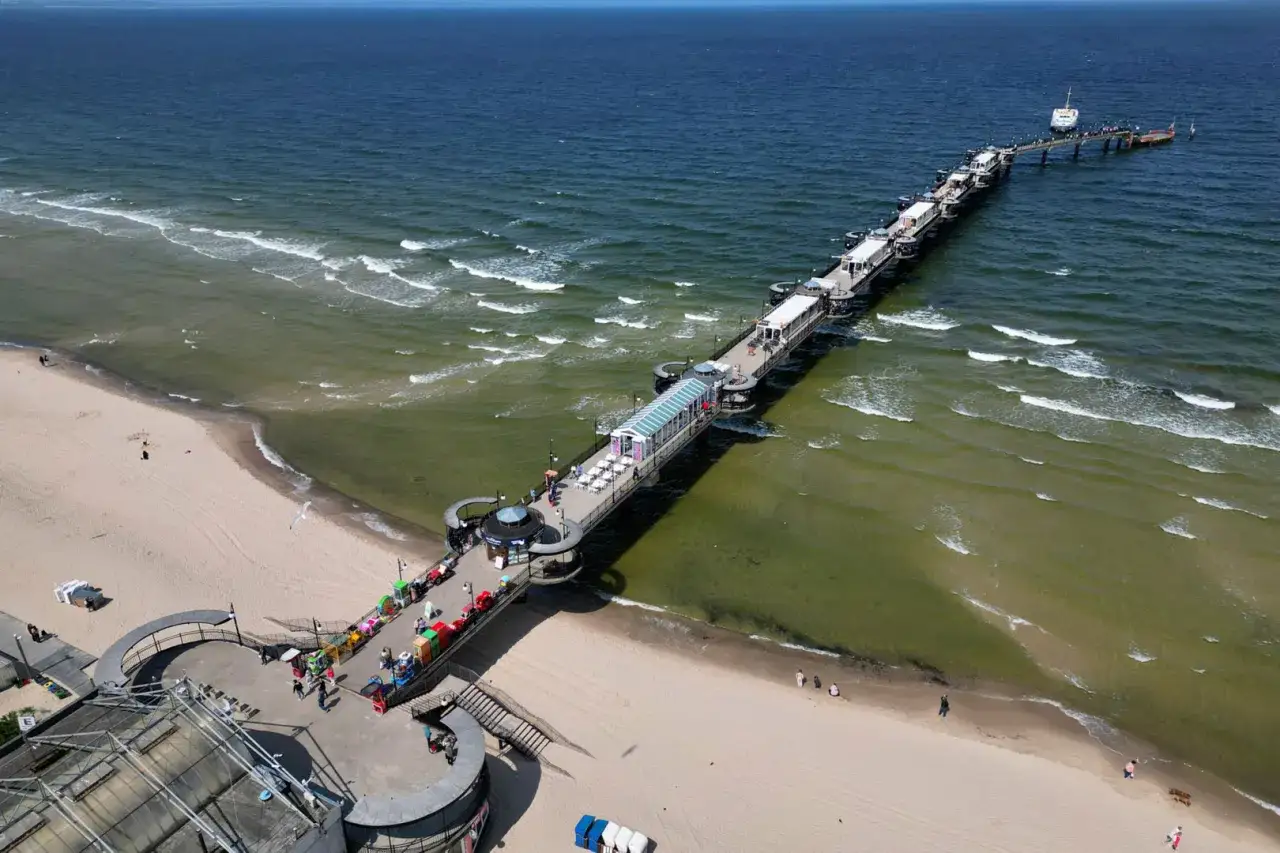Długie molo w Świnoujściu, z kawiarniami i atrakcjami, wchodzi w morze. Spacerowicze cieszą się słońcem na plaży.