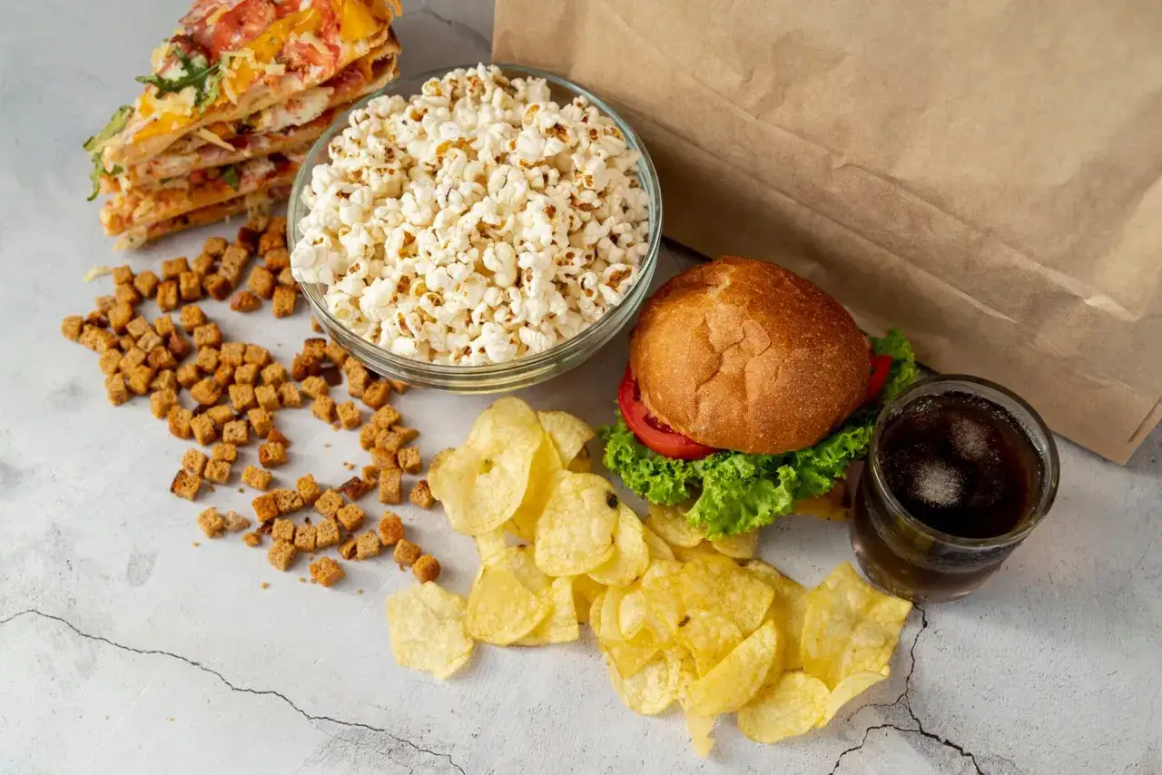 Stos niezdrowych węglowodanów: pizza, popcorn, frytki, grzanki, burger i napój gazowany.