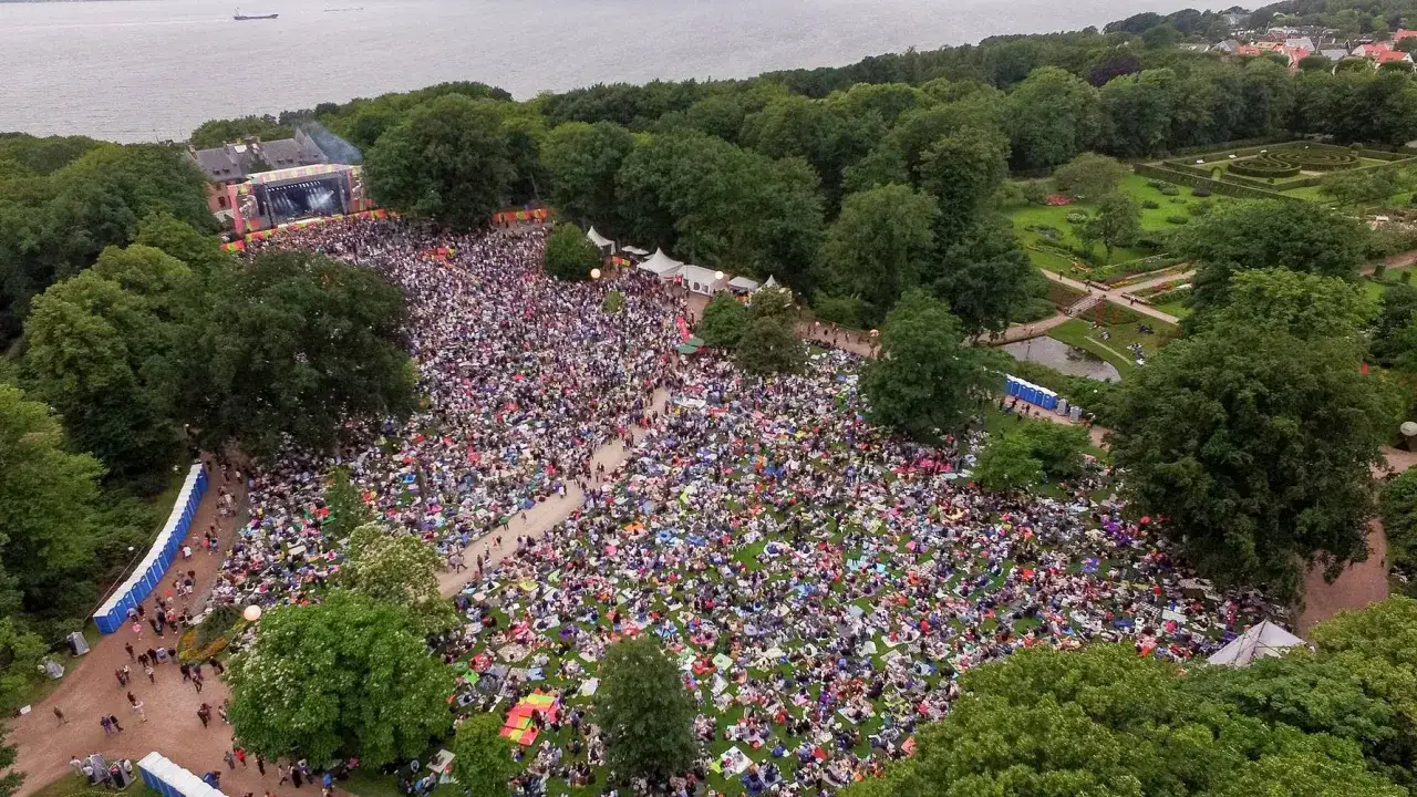 Stor publik njuter av en konsert i botaniska trädgården. Människor sitter på filtar på gräsmattan framför en scen.
