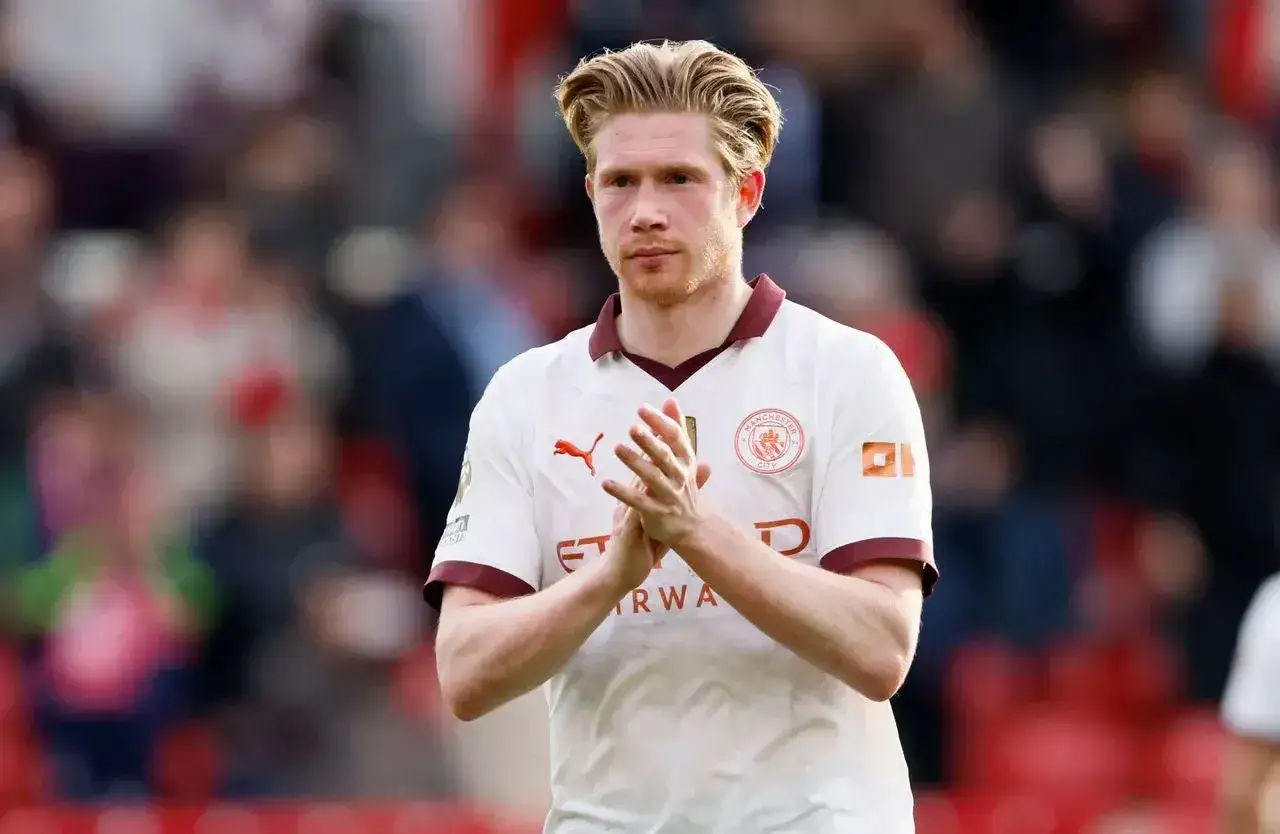 Kevin De Bruyne, w białej koszulce Manchester City, z dłońmi złożonymi w geście uznania.