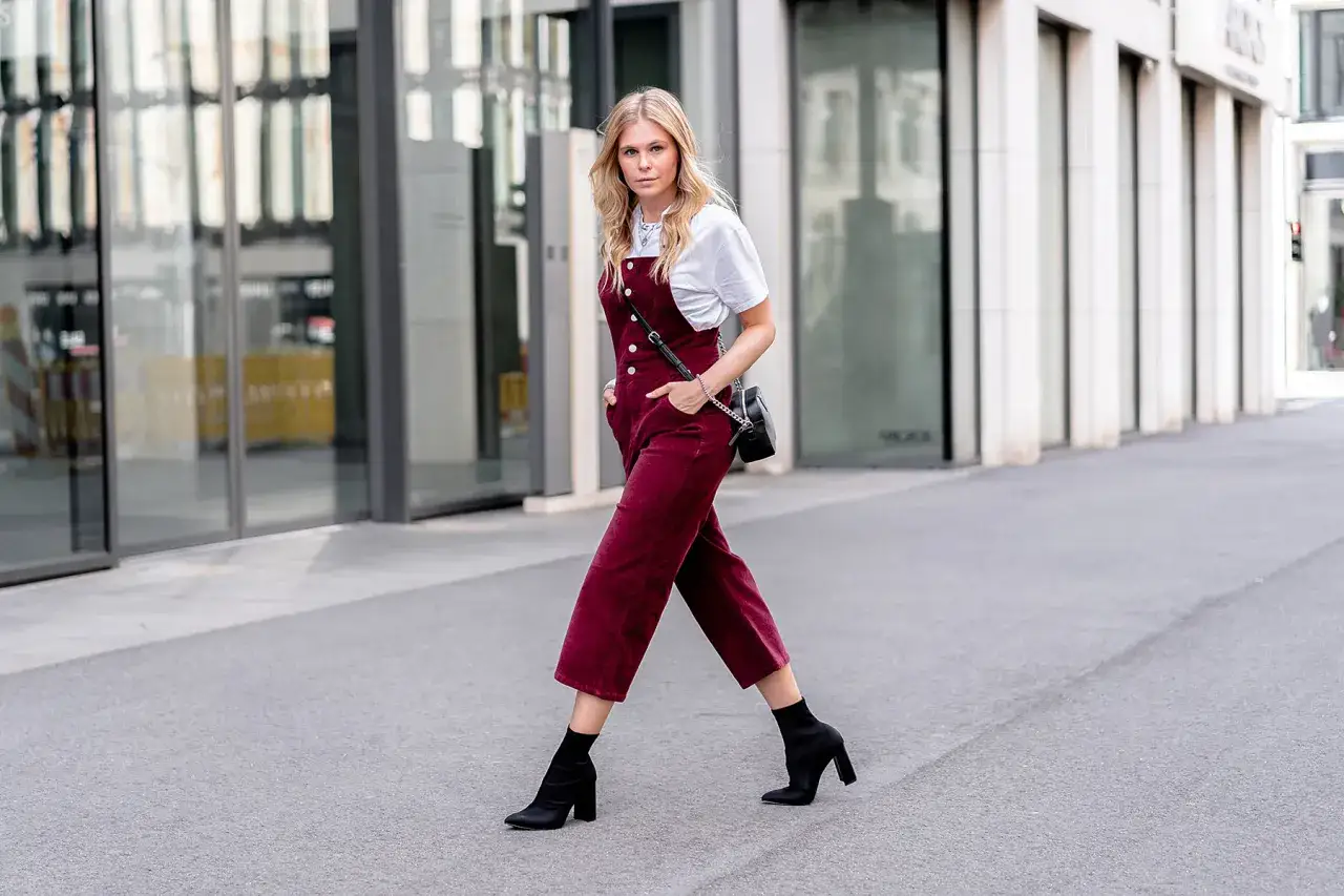 Stylische Frau in bordeauxroter Cordhose mit Latz, kombiniert mit wei&szlig;em T-Shirt und schwarzen Stiefeletten. Ein tolles Beispiel, wie man Cordhose kombinieren Damen kann.