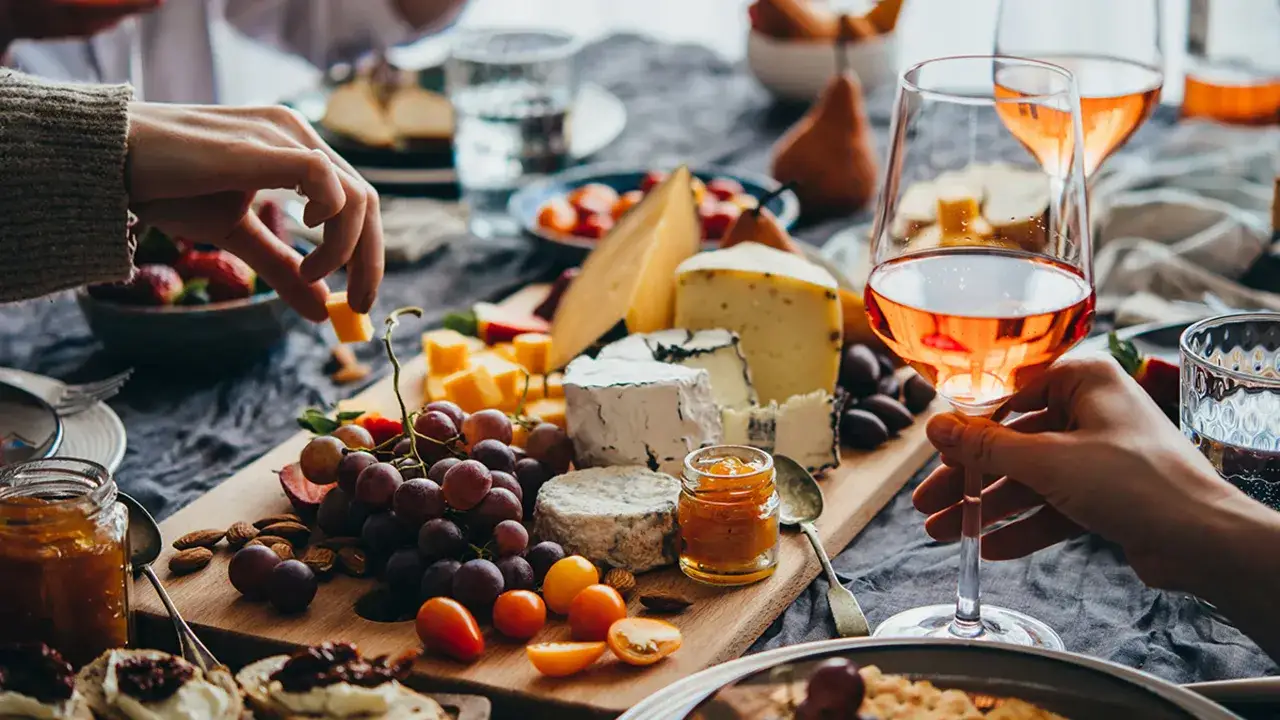 A cheese board with grapes, nuts, and various types of wine, including rosé, is being enjoyed by friends.