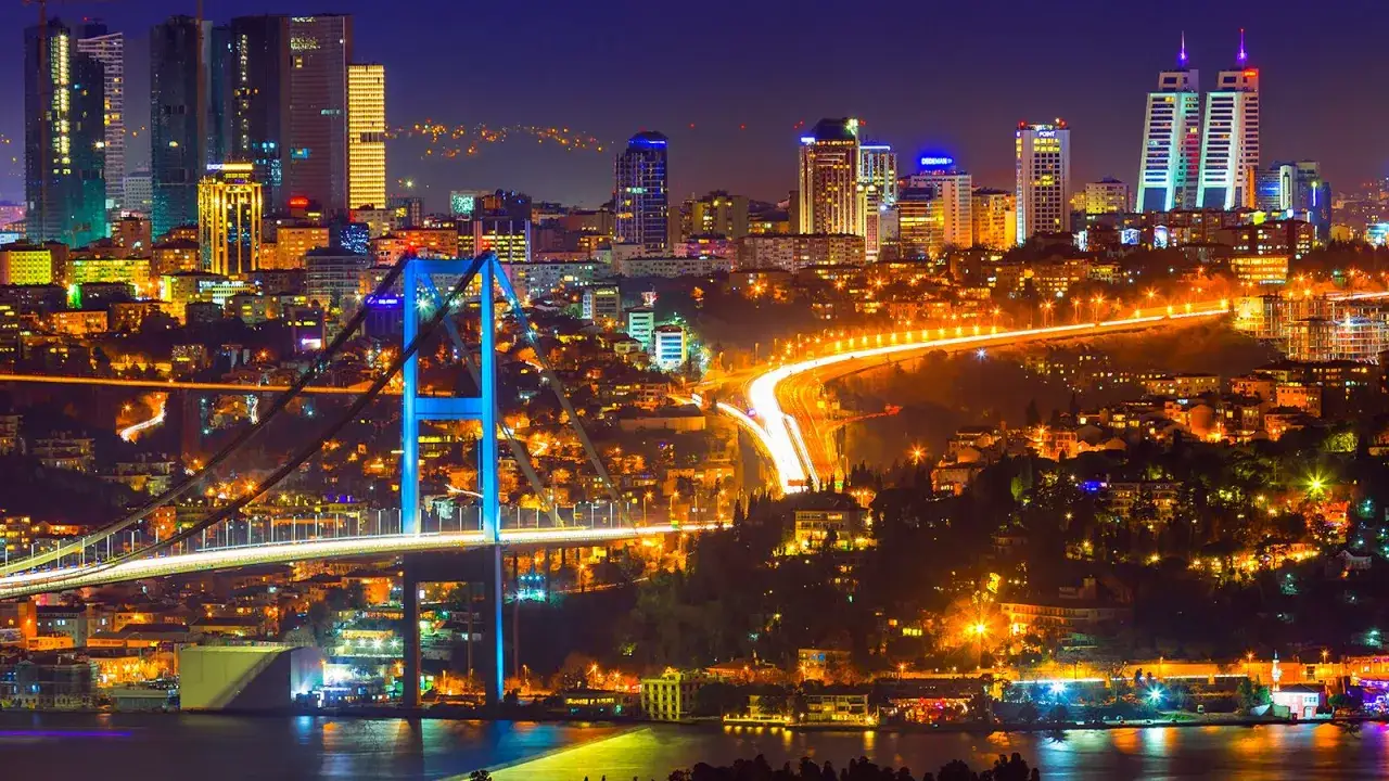 Nocny Stambuł tętni życiem. Most Bosphorus, oświetlony na niebiesko, łączy dwa kontynenty, a światła miasta odzwierciedlają się w wodzie. Stambuł ludność widać w rozświetlonych oknach.