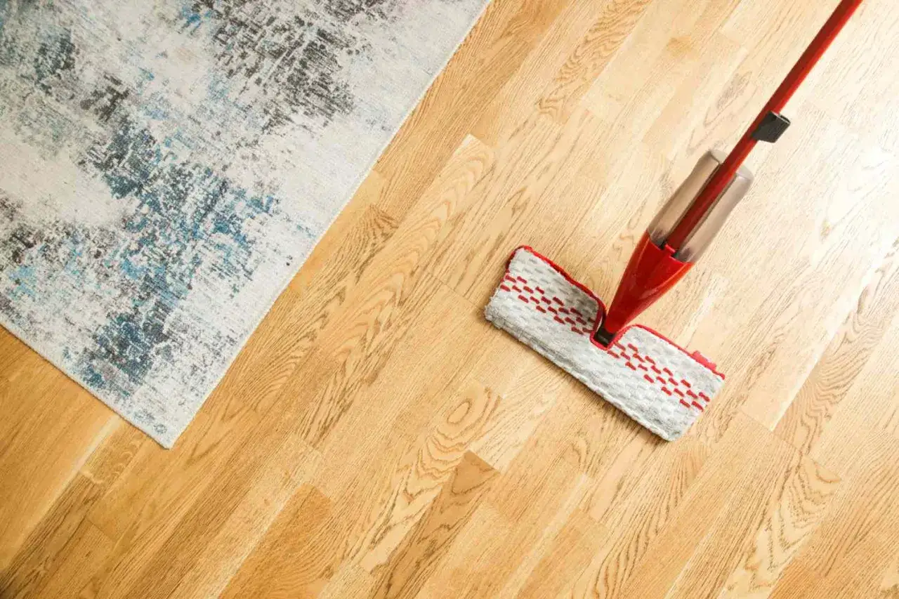 Mopa roja limpiando un suelo de parquet de madera clara, junto a una alfombra con dise&ntilde;o abstracto. Cuidado y mantenimiento del parquet.