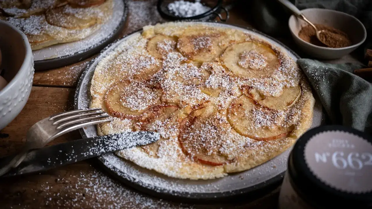 Ein goldbrauner Apfelpfannkuchen, best&auml;ubt mit Puderzucker, wartet darauf, angeschnitten zu werden. Ein k&ouml;stliches Rezept f&uuml;r gem&uuml;tliche Stunden.