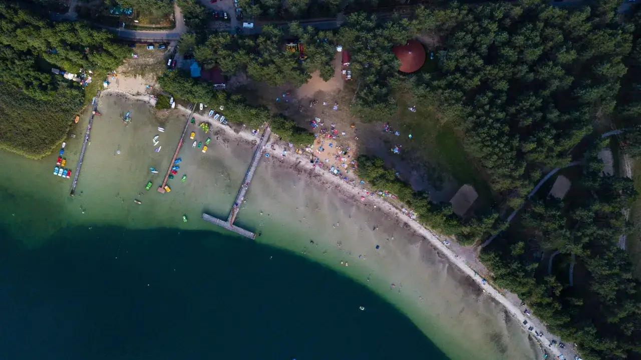 Widok z lotu ptaka na plażę i kąpielisko nad jeziorem Świętajno. Ludzie odpoczywają na piasku, pływają i korzystają z łódek.