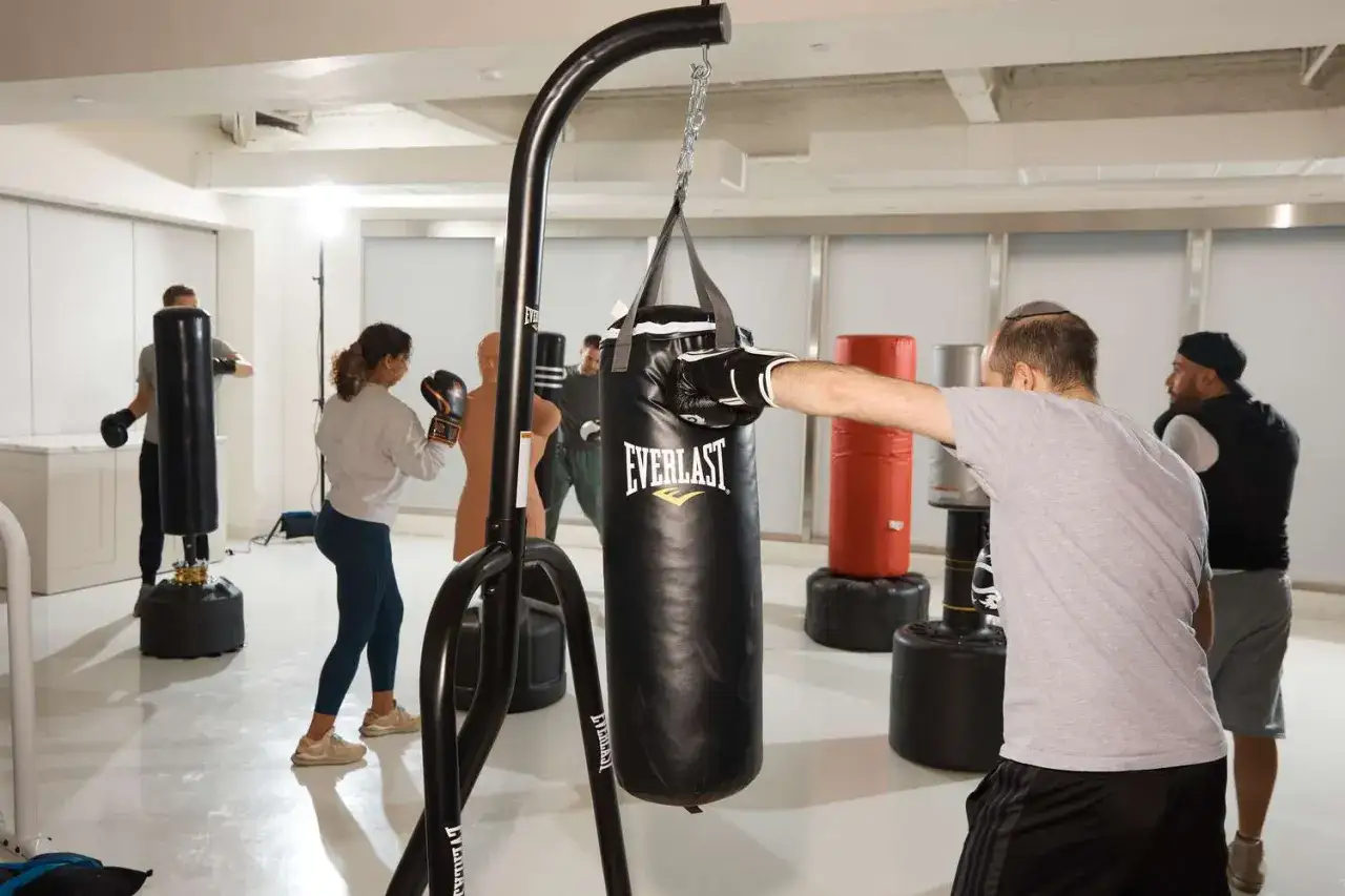People are engaged in boxing workouts at home with bags. A man in a grey shirt throws a punch at a black Everlast bag.