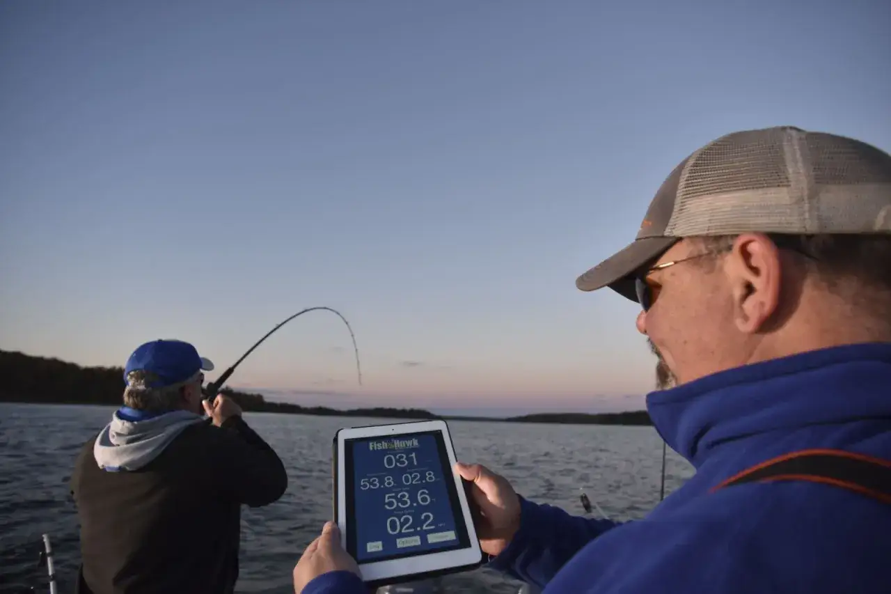 Zwei Angler auf einem Boot, einer wirft die Angel aus, der andere hält ein Tablet mit Fischdaten. Tieferer Start Erfahrungen mit moderner Technik.