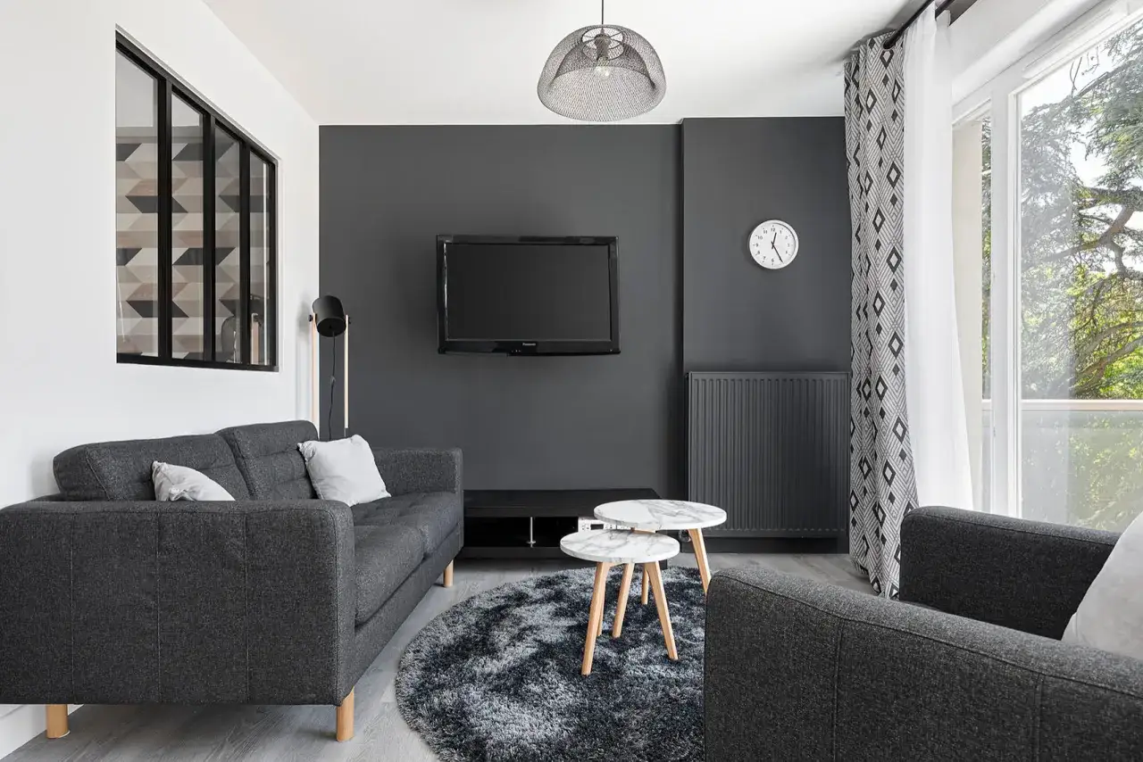 Un petit espace chambre salon moderne avec canapé gris, table basse marbre, tapis rond et TV murale.