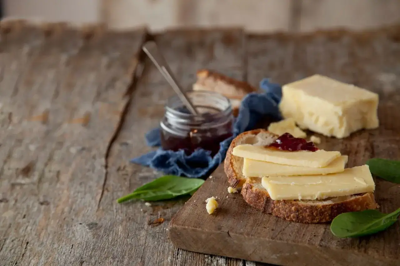 Ein Brot mit Scheiben von mildem Cheddar und Marmelade. Daneben ein Glas Marmelade und ein Stück Cheddar.