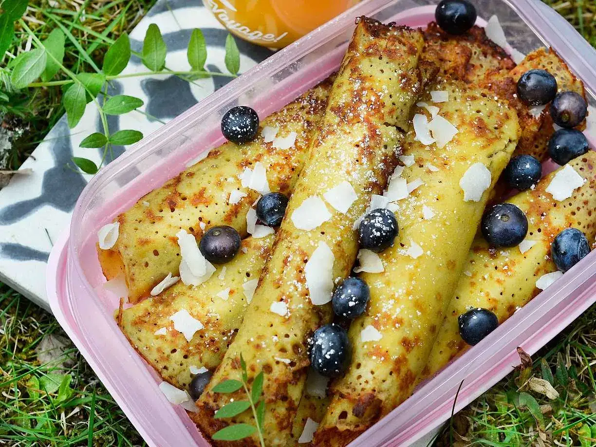 Glutenfria pannkakor rullade och toppade med blåbär och kokosflingor i en rosa matlåda, redo för picknick.