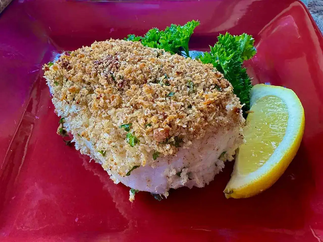 Ein saftiges Fischfilet mit knuspriger Parmesankruste, garniert mit Petersilie und einer Zitronenspalte auf rotem Teller.