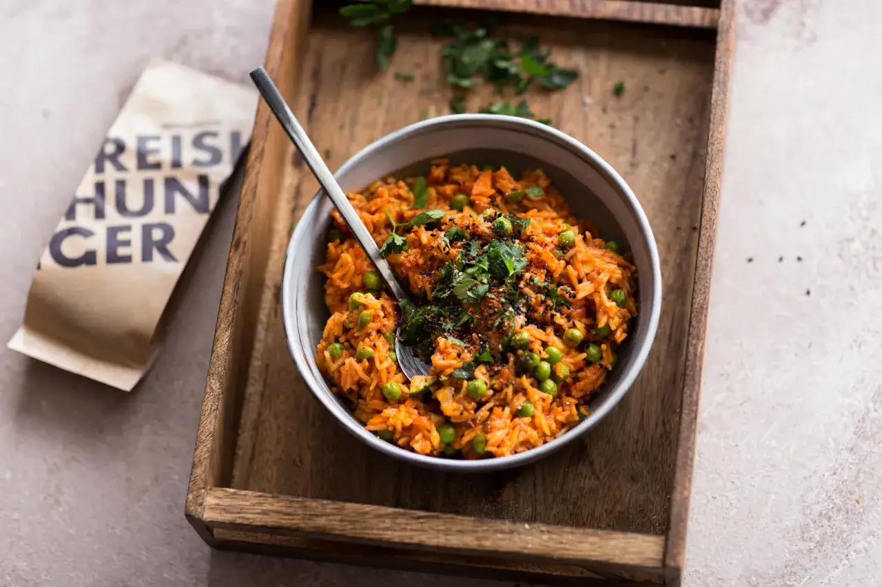 Schüssel mit veganem Djuvec Reis, Erbsen und Karotten, garniert mit Kräutern und Gewürzen. Daneben eine Packung 