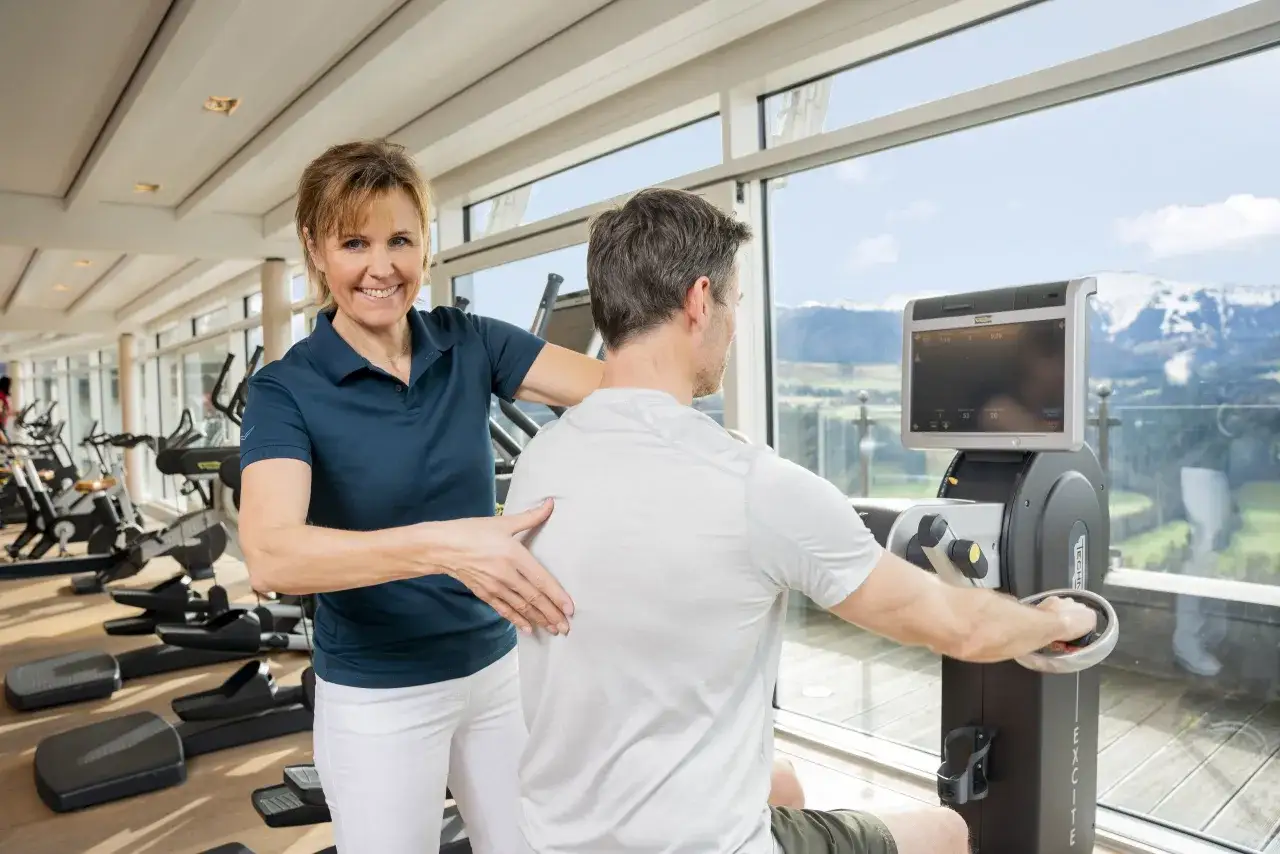 Eine Physiotherapeutin unterstützt einen Mann beim Training an einem Gerät mit Blick auf die Berge.