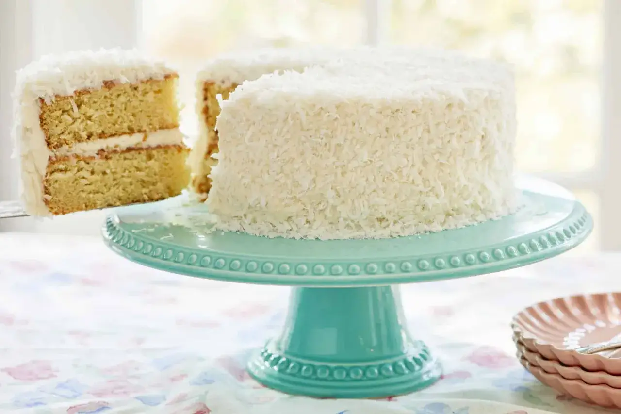 Ein saftiger Buttermilch Kokos Kuchen, bedeckt mit Kokosraspeln, wird auf einem türkisfarbenen Tortenständer präsentiert. Ein Stück wird gerade herausgeschnitten.