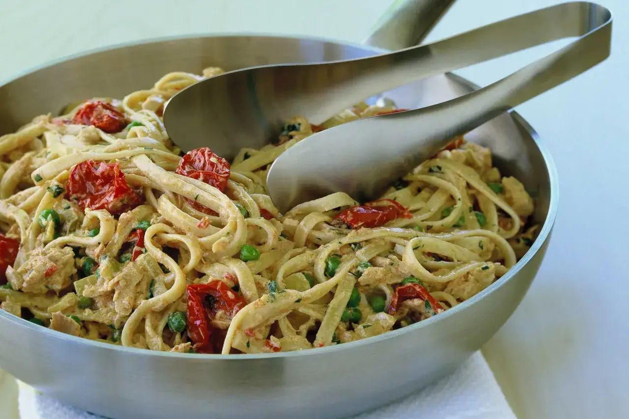 Un plat généreux de pâtes au thon, tomates séchées et petits pois, prêt à être servi avec des pinces.