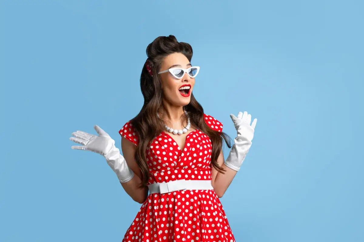 Kobieta w stylu pin-up w czerwonej sukience w grochy, białych rękawiczkach i okularach, z uniesionymi rękami, wyraża radość.