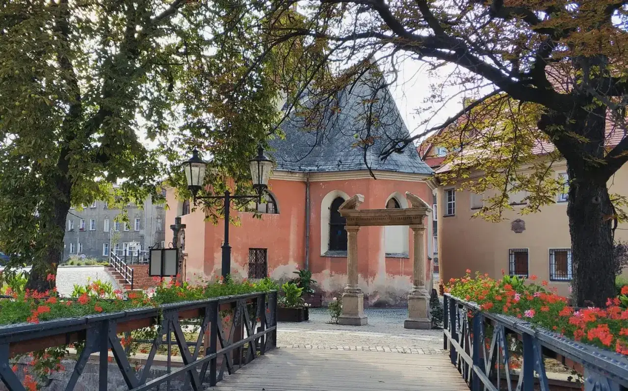 Mostek z kwiatami prowadzi do urokliwego zakątka, gdzie r&oacute;żowa kapliczka i zabytkowe budynki tworzą piękne nowa ruda atrakcje.