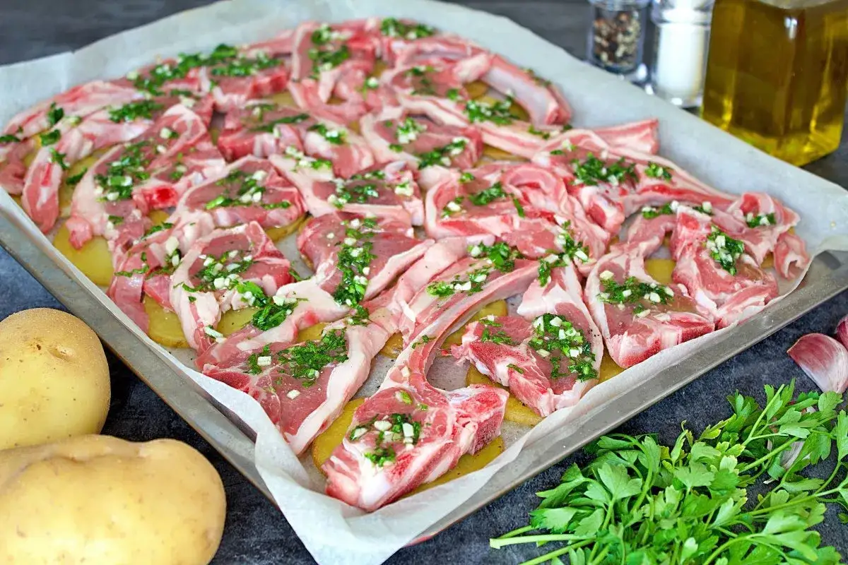 Chuletas de cordero al horno listas para cocinar, con patatas y hierbas frescas.