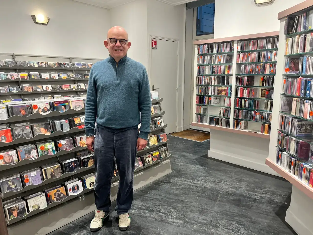 Un homme chauve souriant se tient dans un magasin de disques rempli de CD. Le prix moyen d'un CD de musique varie, mais ces &eacute;tag&egrave;res en regorgent.
