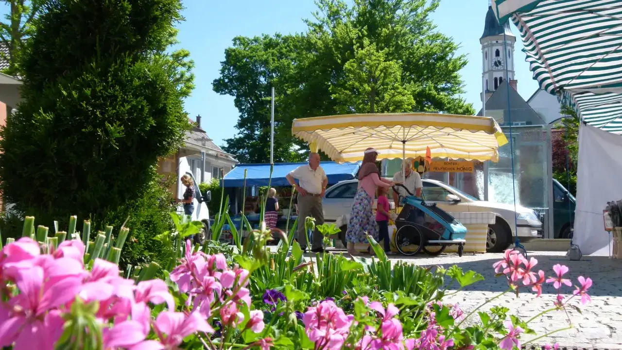 Bunte Blumen blühen am Markt Mering. Menschen kaufen Honig unter Sonnenschirmen, ein Kirchturm ragt empor.