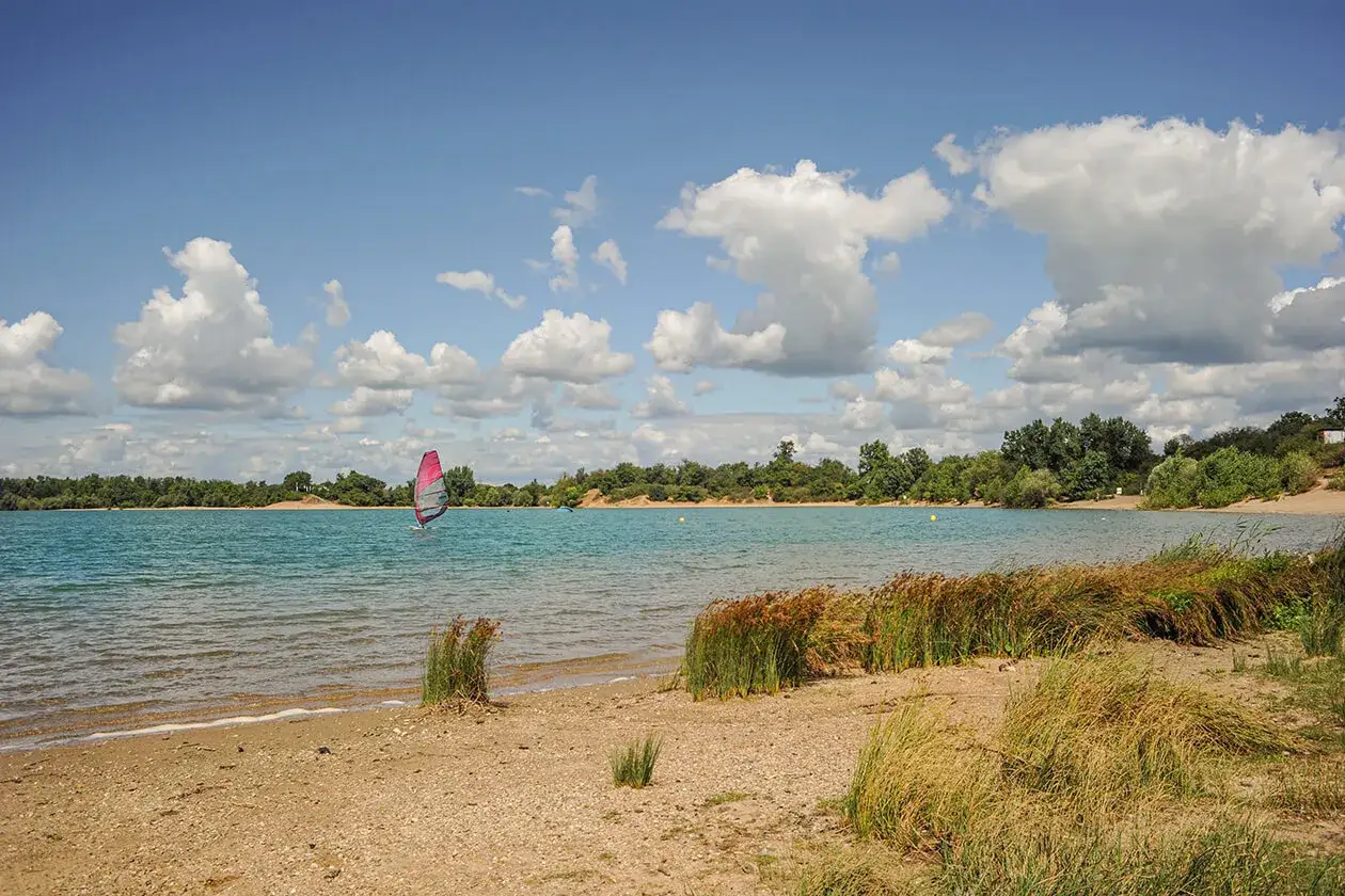 Windsurfer auf dem See bei Karlsruhe, umgeben von gr&uuml;nen Ufern und bew&ouml;lktem Himmel.