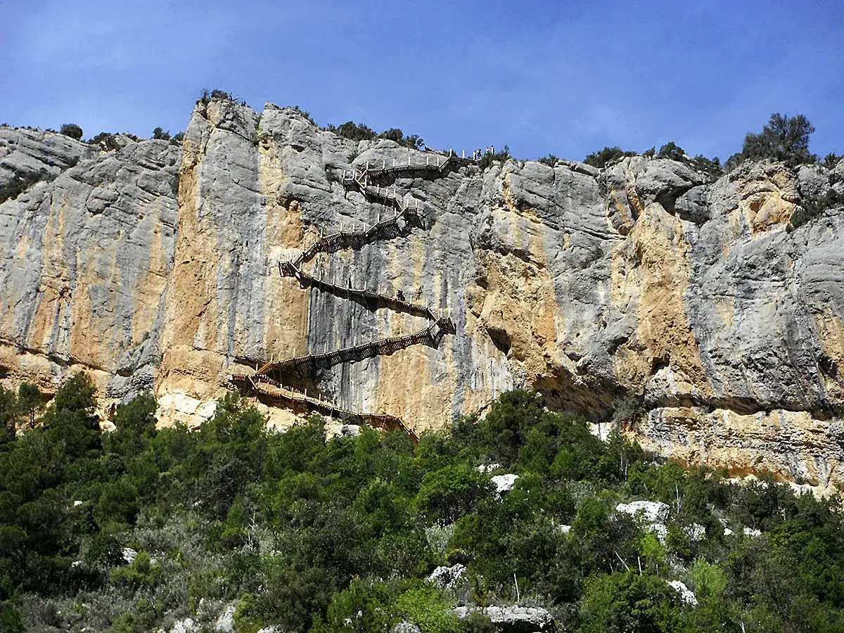 Impresionantes escaleras de Montfalcó serpentean por un acantilado rocoso, rodeadas de vegetación verde.