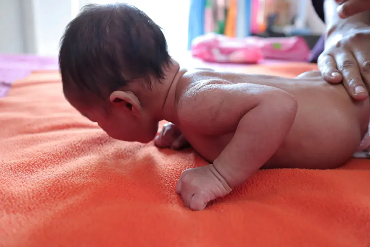 Un beb&eacute; boca abajo sobre una manta naranja, con una mano adulta sujetando su espalda. Es importante saber porque hay que sujetar la cabeza a los beb&eacute;s.