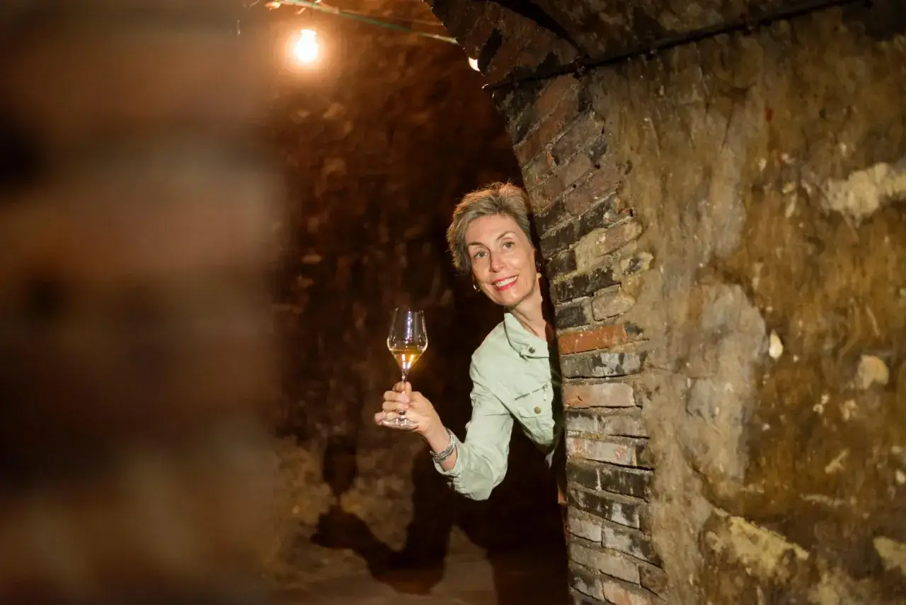 Une femme souriante tient un verre de vin clair dans une cave vo&ucirc;t&eacute;e en briques.