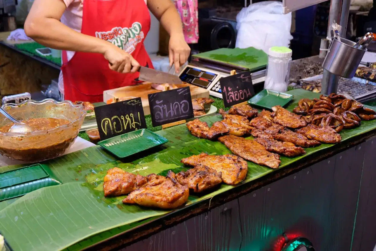 Grillad mat p&aring; bananblad, en del av den rika **thailand matkultur**. En s&auml;ljare sk&auml;r upp k&ouml;tt.