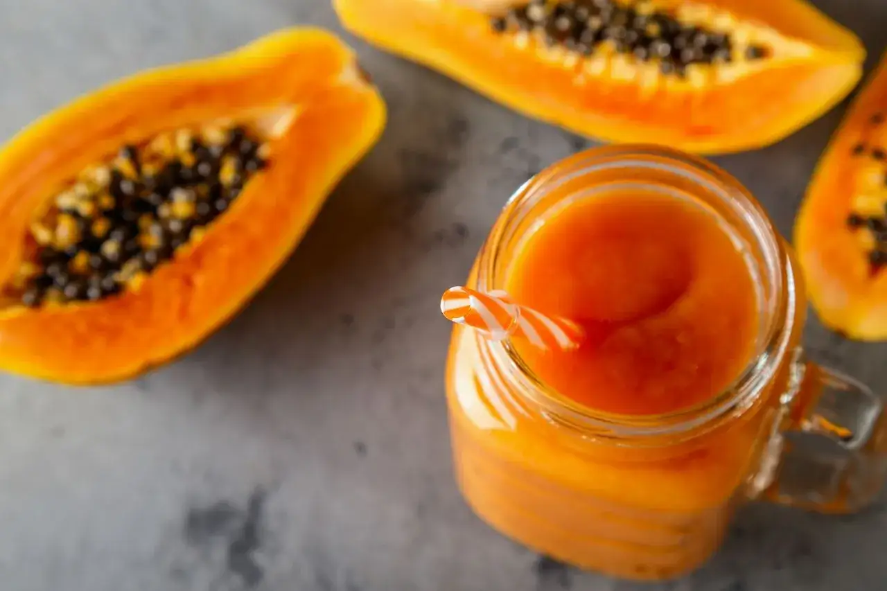 Frullato di papaya in un bicchiere con cannuccia a righe, accanto a metà papaya. Un aiuto naturale per chi vuole imparare come sgonfiare la pancia.