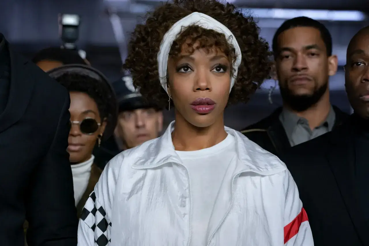 Młoda kobieta w białej opasce i sportowej kurtce, przypominająca Whitney Houston z filmu, otoczona przez ludzi.