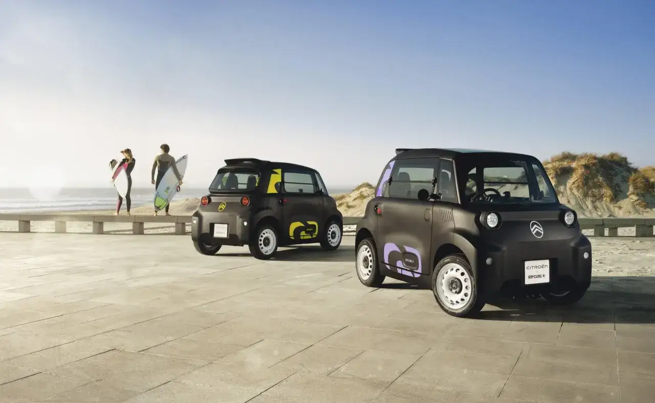 Dwa czarne Citroen Ami Buggy z kolorowymi akcentami na plaży, obok surfer&oacute;w.
