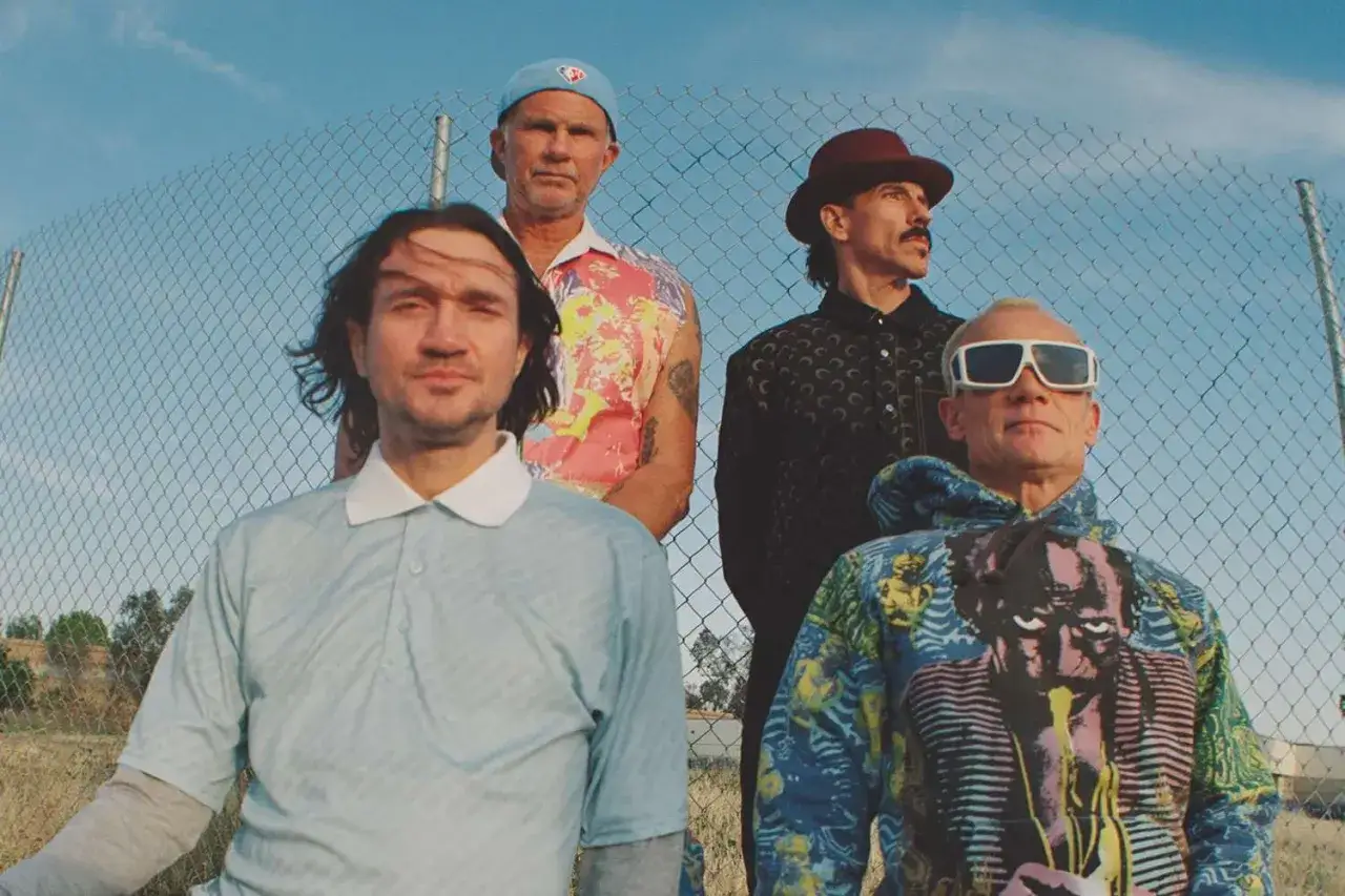 Les membres des Red Hot Chili Peppers posent devant une clôture. Flea porte des lunettes de soleil et un sweat à capuche coloré.