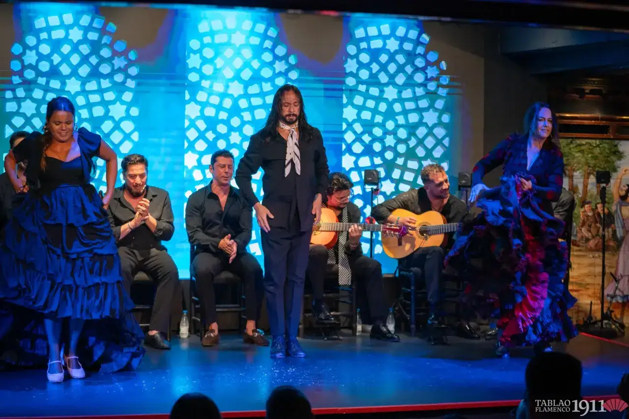 Artistas de flamenco espa&ntilde;ol en escena. Una bailarina con un vestido azul y otra con uno rojo y negro, acompa&ntilde;adas por guitarristas y un hombre con pa&ntilde;uelo.