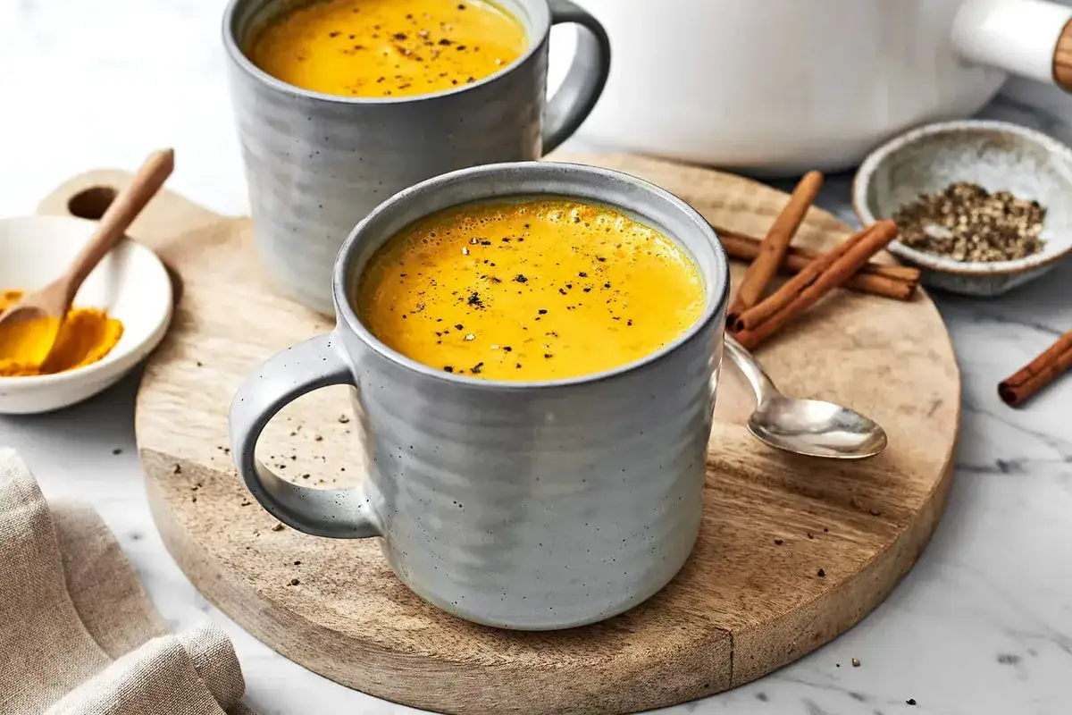 Zwei Tassen goldene Milch, bereit, ihre wohltuende goldene Milch Wirkung zu entfalten. Zimtstangen und Kurkumapulver liegen daneben.