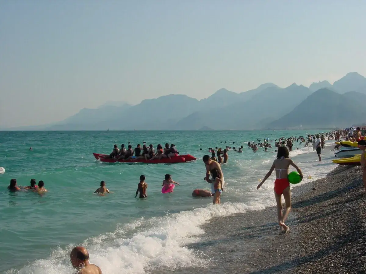 Menschen genießen einen sonnigen Tag am Antalya Strand. Kinder planschen im Meer, während andere auf einer Bananenboot-Fahrt Spaß haben. Im Hintergrund erheben sich majestätische Berge.
