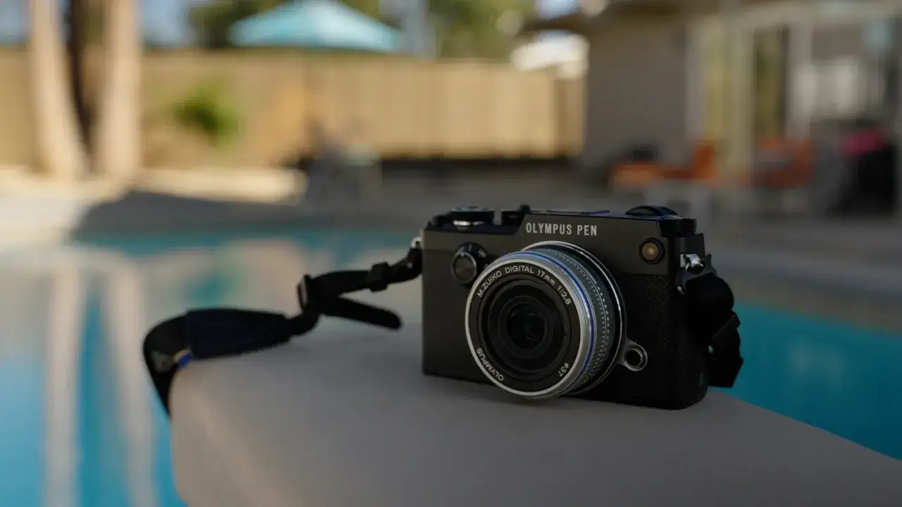 An Olympus PEN-F camera rests on a ledge by a pool, ready to capture summer memories.