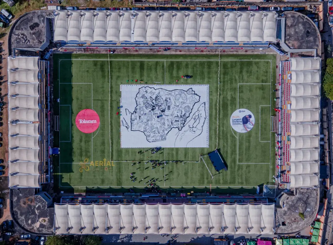 Luftaufnahme eines Stadions mit dem größten Gemälde der Welt auf dem Spielfeld. Menschen stehen auf dem Spielfeld und betrachten das Kunstwerk.