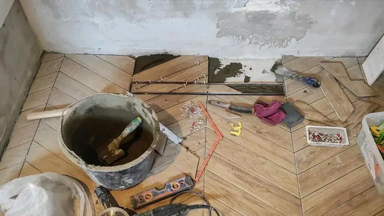 Układanie drewnopodobnych płytek na płytki w jodełkę. Widać wiadro z zaprawą, kielnię, poziomice i narzędzia.