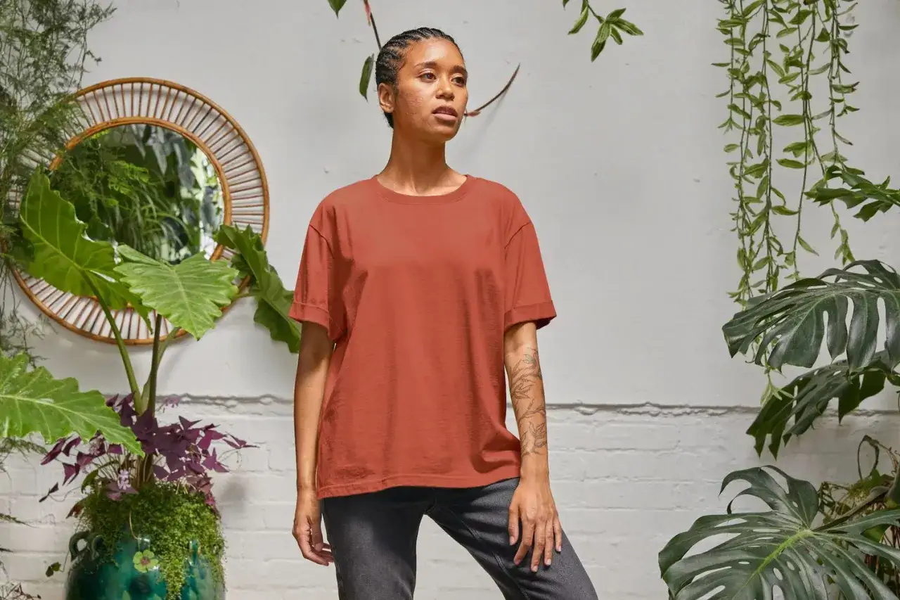 Koszula oversize damska w kolorze ceglanym, idealna do casualowych stylizacji. Modelka w luźnym t-shircie i szarych spodniach, otoczona bujną zielenią.