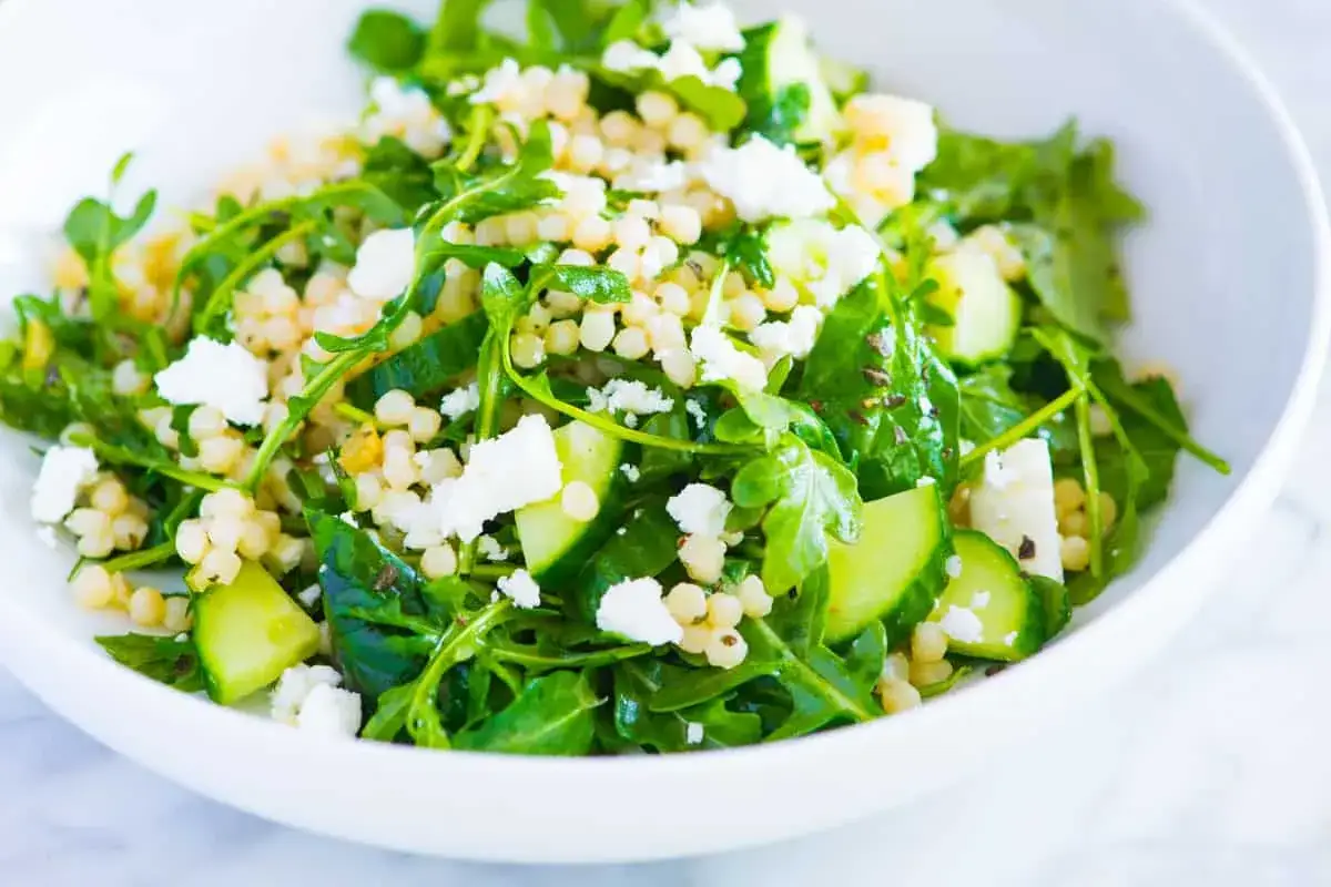 Frischer Couscous Salat mit Gurkenst&uuml;cken und zerbr&ouml;seltem Feta in einer wei&szlig;en Sch&uuml;ssel.