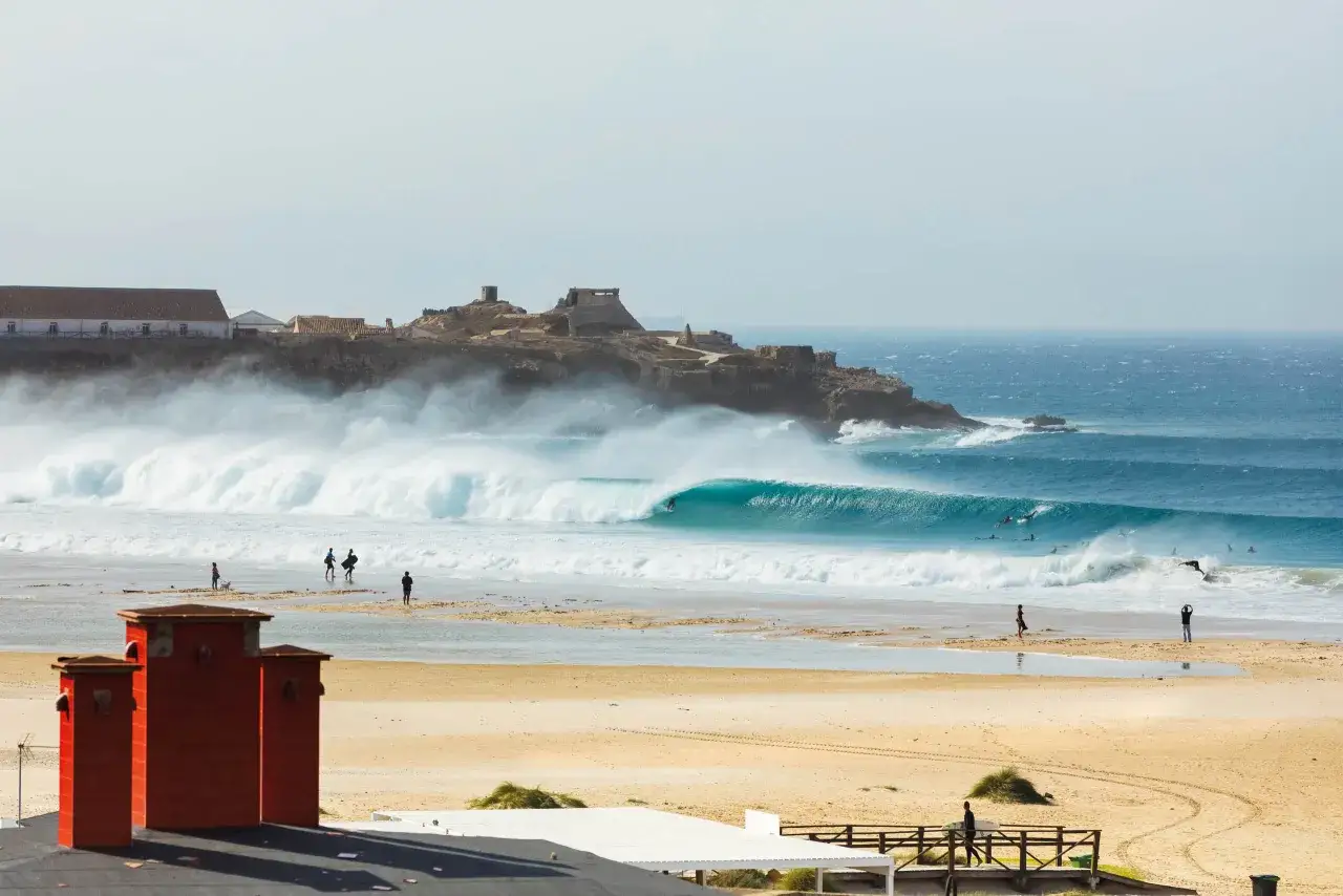 Perfekte Wellen f&uuml;r **Tarifa Surfen**. Surfer reiten die t&uuml;rkisfarbene Welle vor einer felsigen K&uuml;ste mit Ruinen.