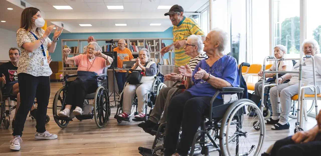 Un gruppo di anziani in sedie a rotelle partecipa a un'attivit&agrave; in una struttura convenzionata per malati di Alzheimer, guidati da un'animatrice.
