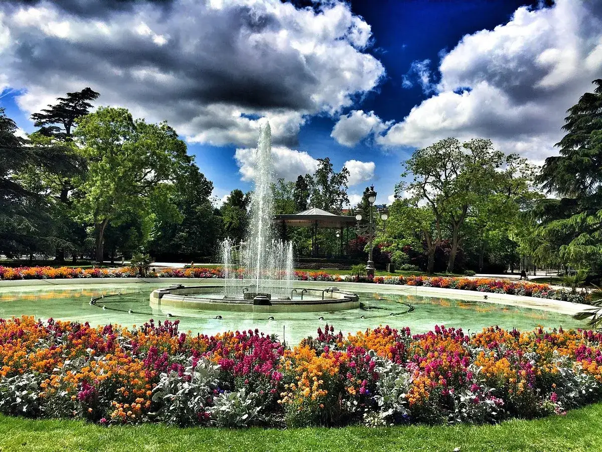 Fontaine d'eau jaillissante au jardin des plantes Toulouse, entourée de fleurs colorées et d'arbres verdoyants sous un ciel nuageux.