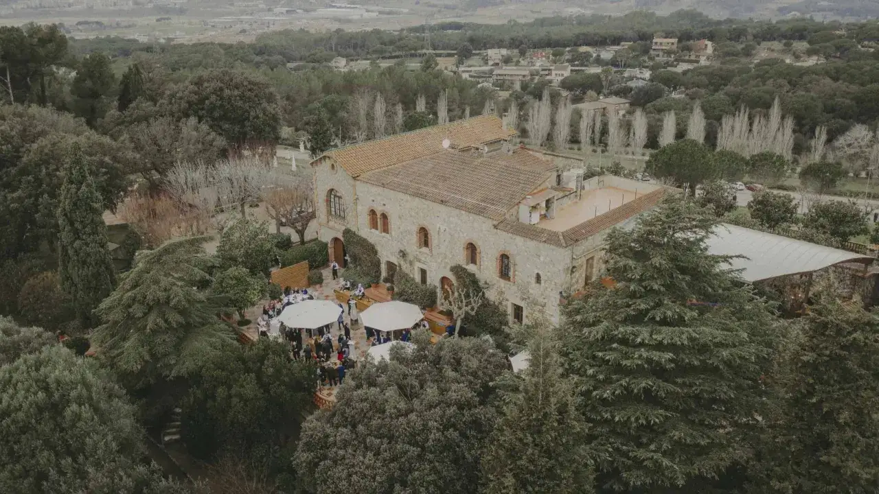 Vista a&eacute;rea de una **boda &iacute;ntima** en un patio rodeado de &aacute;rboles. Invitados reunidos bajo carpas blancas frente a una casa de piedra.