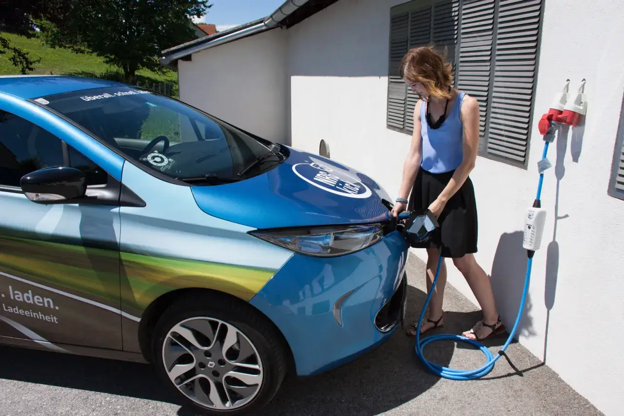 Kobieta podłącza niebieską hybrydę plug-in do ładowania przy białej ścianie.